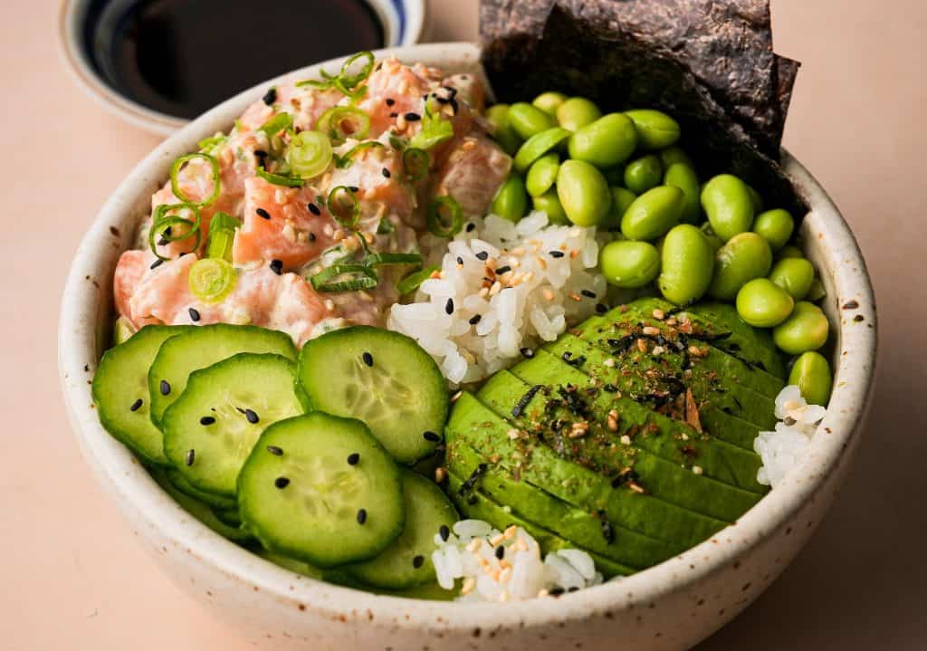 salmon poke bowl garnished with sliced avocado, cucumber, and edamame beside a small saucer of ponzu sauce